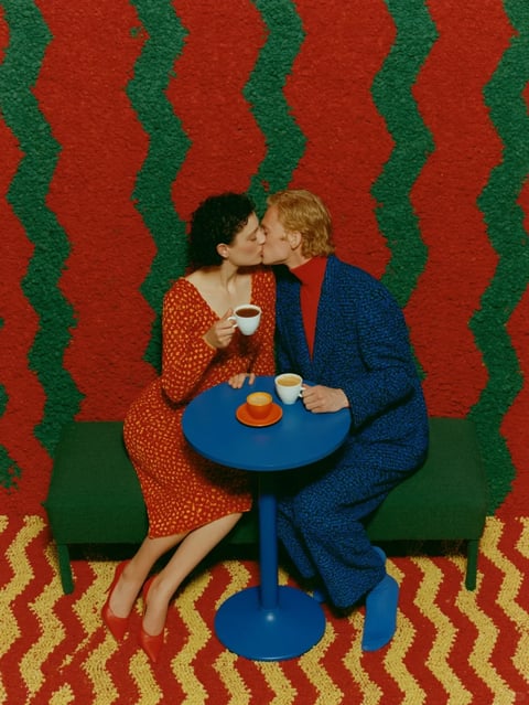 The image is a vertically oriented, photorealistic studio photograph featuring a couple seated on a low green bench, sharing a kiss over a small blue round table. The scene is dominated by a vibrant, high-saturation color palette of primary red, cobalt blue, and forest green, with accents of orange and yellow. The background consists of a textured, shag-carpet-like wall with a large, repeating zigzag pattern in deep red and forest green, creating a strong graphic and textural backdrop. The floor mirrors this pattern but in red and yellow. The woman on the left wears a red dress with a small yellow floral pattern and red heels, holding a white cup. The man on the right wears a textured blue suit with a red turtleneck and bright blue boots, holding another white cup. A third orange cup sits on a matching saucer on the table between them. The lighting is bright, even, and frontal, casting minimal soft shadows, which flattens the space and enhances the graphic, color-blocked aesthetic. The composition is centered and symmetrical, with the couple and table forming the focal point against the repetitive background. The overall visual style is highly stylized, retro-inspired (evoking 1970s pop art and interior design), with a bold emphasis on saturated color fields, geometric shapes (zigzags, circles), and contrasting textures between the fuzzy carpet and smooth table/cups. There is no visible text in the image.