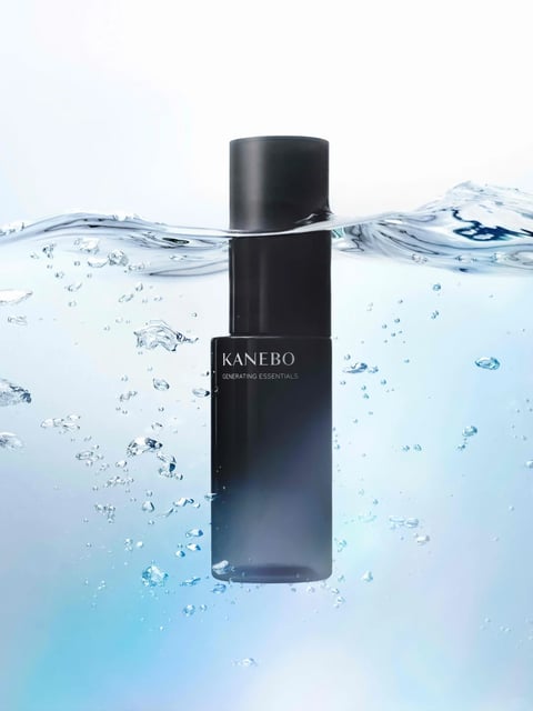 The image features a sleek, black cylindrical bottle of KANEBO GENERATING ESSENTIALS, partially submerged in clear, dynamic water. The composition is centered, with the bottle vertically oriented, its top half above the waterline and the lower half immersed, surrounded by rising air bubbles and gentle ripples on the water's surface, set against a soft, gradient light blue to white background. Visible text appears on the bottle: the brand name "KANEBO" in a clean, sans-serif font, with "GENERATING ESSENTIALS" in a smaller, similar typeface directly below it, both in white against the dark bottle, serving as clear product identification. The visual aesthetic is photorealistic, emphasizing high-end product photography with a clean, minimalist style and precise rendering. The color language is dominated by cool tones, with a high-contrast palette of deep black against clear water and a light blue-white gradient, creating a crisp, modern feel. Lighting is soft and diffused, appearing to come from behind or above, highlighting the bottle's form and the translucency of the water while casting subtle shadows for depth. Texture and material feel are distinctly smooth and glossy on the bottle's surface, contrasted with the liquid, bubbly, and refractive qualities of the water. The composition uses a centered, symmetrical framing that draws focus directly to the product, with the waterline creating a horizontal division that adds dynamic interest and depth. Shape language is primarily geometric, with the cylindrical bottle's clean, straight lines and defined edges, juxtaposed against the organic, fluid forms of the water and bubbles. The mood and atmosphere convey a sense of purity, sophistication, and freshness, with an energetic yet calm tone from the water's movement, suggesting hydration and premium quality.