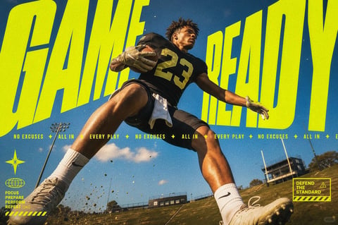 The image is a dynamic, high-energy sports graphic featuring a male American football player as the central subject, captured mid-action on a grassy field under a bright, partly cloudy blue sky. The composition uses a dramatic low-angle, wide-angle perspective that elongates the athlete's limbs and creates a sense of power and forward motion, with dirt and grass kicked up near his cleats. The player, wearing a black jersey with the number 23, black shorts, and white Nike socks and cleats, is in a running or leaping stance, holding a football securely in his left arm while his right arm is extended for balance. Dominating the background is massive, bold, lime-yellow typography spelling "GAME READY," which arcs behind the player and integrates with the sky, creating a layered, collage-like effect. A repeating ticker-tape style text strip runs horizontally across the middle, reading "NO EXCUSES + ALL IN + EVERY PLAY + NO EXCUSES + ALL IN + EVERY PLAY + NO EXCUSES + ALL IN + E..." in the same lime-yellow. Additional smaller graphic elements include a star icon, a globe icon with the stacked text "FOCUS PREPARE PERFORM REPEAT 24/7," and a yellow caution-tape style box in the lower right with the text "DEFEND THE STANDARD" and a warning symbol. The visual aesthetic blends photorealistic elements with graphic design, characterized by high contrast, saturated colors, and a slightly gritty, textured overlay that adds a tactile, worn feel. The lighting is bright and directional, suggesting midday sun, casting sharp shadows and highlighting the player's musculature with warm tones against the cool blue sky. The color palette is dominated by the vibrant lime-yellow of the text, the deep blue of the sky, the earthy greens and browns of the field, and the black and white of the uniform, creating a high-contrast, energetic visual language. The mood is motivational, intense, and focused, emphasizing athletic readiness and determination through its composition, color, and text.