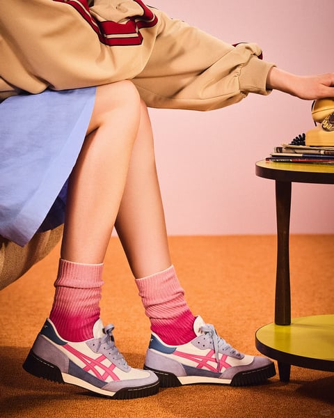 The image captures a stylized, photorealistic close-up of a person's lower body seated on a textured surface, with a focus on fashion details against a retro-inspired backdrop. The composition centers on the subject's legs, angled slightly, showcasing a layered outfit: a beige sweatshirt with red and navy striped cuffs and hem, a light blue skirt, vibrant pink ribbed socks, and multi-colored sneakers featuring white, lavender, pink, and navy panels with a distinctive crisscross pattern. To the right, a small, round table with a yellow top and dark legs holds a stack of books and a vintage yellow rotary phone, which the subject's hand is reaching for. The color palette is saturated and warm, dominated by soft pinks, muted blues, and earthy oranges, with high contrast between the pastel tones of the clothing and the richer hues of the floor and background. Lighting is soft and directional from the left, casting gentle shadows that emphasize the textures of the knit socks, suede-like shoe panels, and woven fabric of the skirt. The visual aesthetic is polished and editorial, reminiscent of retro fashion photography with a clean, focused composition that highlights material textures and color blocking. There is no obvious text visible in the image; the focus is entirely on the tactile quality of the garments, the playful color coordination, and the nostalgic atmosphere created by the vintage props and warm, glowing light.