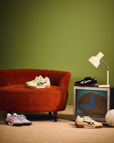 The image is a photorealistic, retro-styled still life composition showcasing four pairs of Onitsuka Tiger sneakers in a domestic setting, with a cohesive mid-century modern aesthetic. The scene is arranged against a flat, matte olive-green wall, with a plush, rust-orange corduroy armchair on the left and a vintage CRT television acting as a pedestal on the right, topped with a single black-and-white sneaker and a classic white gooseneck desk lamp. The lighting is soft and diffused, appearing to come from the upper left, casting gentle, elongated shadows that enhance the three-dimensionality of the objects and create a warm, nostalgic atmosphere. The color palette is muted and earthy, dominated by the olive-green background, rust-orange chair, and beige carpet, with the sneakers providing accent colors in pastel mint, lavender, and classic black-and-white. Textures are richly rendered, from the deep, tactile ribs of the corduroy upholstery to the smooth, matte finish of the sneaker uppers and the slightly reflective screen of the television, which displays a close-up graphic of a sneaker's gum sole. The composition is balanced and slightly asymmetrical, using the low angle and the placement of shoes on the floor, chair, and TV to create depth and guide the eye through the scene. The shapes are a mix of the chair's soft, organic curves and the geometric, clean lines of the footwear and electronics. The overall mood is calm, curated, and stylistically nostalgic, evoking a comfortable, vintage-inspired interior.
