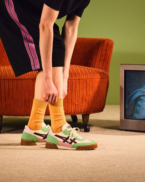 The image presents a photorealistic, stylistically focused shot of a person's lower legs and feet, centered in a retro-inspired interior scene. The primary subject is a person seated on an orange corduroy armchair, leaning forward to adjust or display a pair of colorful athletic shoes. The shoes feature a cream base with panels in mint green, soft pink, and black, complemented by a thick, ribbed brown sole. They are worn with mustard-yellow ribbed crew socks. The person's legs are clad in black track pants with a prominent vertical stripe in purple and pink along the outer seam. The scene is set on a beige carpet against a muted, olive-green wall. To the right, a portion of a vintage television set is visible, displaying a blue-toned image of a fabric bag. The lighting is soft and diffused, casting gentle shadows and creating a warm, inviting atmosphere with a focus on texture and color harmony. There is no obvious readable text in the image.