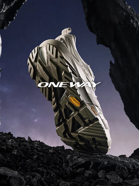 The image presents a low-angle, photorealistic shot of a single light-colored trail running shoe, angled to prominently display its aggressive, multi-directional tread pattern and Vibram-branded sole. The shoe is positioned as if stepping onto a pile of dark, jagged volcanic rocks in the foreground, with larger, silhouetted rock formations framing the top corners against a deep twilight sky that gradients from dark blue to a soft purple haze. The composition uses dramatic perspective and framing to emphasize the shoe's rugged, technical design and its readiness for challenging terrain. The visual aesthetic is sleek and high-tech, with a focus on material realism, showcasing the shoe's layered synthetic upper, mesh details, and the complex geometric shapes of the rubber outsole. The color palette is cool and desaturated, dominated by slate blues, purples, and grays, with the light bone-colored shoe providing contrast and a small, warm orange Vibram logo offering a focal point. Lighting is directional and moody, coming from the upper left, casting hard shadows that accentuate the textures of both the shoe's tread and the rocky surfaces, creating a sense of dramatic anticipation and performance-oriented capability. Overlaid on the image is the text "ONE WAY" in a bold, white, stylized sans-serif font, positioned across the midsection of the shoe; this text acts as a brand identifier or slogan, reinforcing the focused, determined theme of the visual.