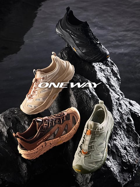 This image presents a dynamic product shot of four athletic shoes arranged on a cluster of dark, wet volcanic rocks, with a backdrop of still, dark water. The composition is energetic and asymmetric, with the shoes positioned at various angles to showcase their profiles, soles, and upper designs. The overall aesthetic is photorealistic and rugged, emphasizing an outdoor, adventure-ready mood. The dominant color palette features earthy, muted tones—black, warm beige, tan with brown accents, and olive green—set against the cool, dark grays of the rocks and water, creating high contrast. Lighting appears to be directional, possibly from the upper left, casting soft yet defined shadows that highlight the textured surfaces of both the shoes and rocks, enhancing their three-dimensional form. Textures are a key element: the rocks are rendered with a glossy, wet, and porous surface, while the shoes display a mix of matte synthetic overlays, breathable mesh panels, and rubberized soles with visible treads and speckled details. The text 'ONE WAY' is prominently overlaid in a bold, white, sans-serif font across the center of the image, serving as a brand identifier. The shape language is a blend of organic (the rugged rocks) and technical geometric (the structured, angular lines of the shoe uppers and outsoles). The overall mood is adventurous, durable, and performance-focused, with a cool, slightly moody atmosphere conveyed by the dark environment and focused lighting.