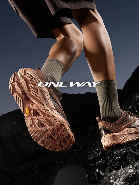 This image presents a dynamic, low-angle close-up of a person's lower legs and feet in mid-stride over dark, wet, rugged rocks, capturing a moment of outdoor action. The composition is dominated by the strong diagonal lines of the legs and the prominent, textured sole of the leading brown trail running shoe, which is angled toward the viewer to showcase its aggressive, geometric tread pattern. A smaller, secondary shoe is visible on the trailing foot. The person wears olive green ribbed socks and dark shorts. The lighting is dramatic and directional, seemingly from the upper left, casting hard shadows that emphasize the muscular definition of the legs and the intricate, gritty texture of the shoe soles and the rocky terrain. The color palette is earthy and high-contrast, featuring dominant tones of warm brown and terracotta in the footwear, muted olive in the socks, and deep charcoal and black in the background rocks, all set against a subtle gradient sky of dark blue to grey. The visual aesthetic is photorealistic with a sharp focus on texture and material, conveying a sense of rugged performance. The text 'ONE WAY' appears twice: once as a large, white, sans-serif graphic overlay centered between the legs, and again in smaller, matching white lettering on the side of the trailing shoe. The overall mood is one of determined energy, physical exertion, and connection with a harsh natural environment, emphasizing the gear's functionality and durability.