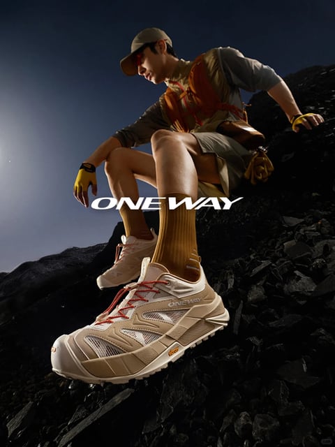 The image is a photorealistic, vertically oriented action-style advertisement featuring a male model in a dynamic climbing pose on a dark, rocky, volcanic terrain. The composition uses a low-angle perspective, placing a large, detailed hiking shoe in the foreground as the primary focal point, with the model's body receding into the mid-ground and a deep blue, gradient sky in the background. The model, dressed in a layered outfit of khaki, olive, and grey with a tan cap, is captured in a crouched, stable stance, gripping the rocks. The color language is dominated by a high-contrast palette of earthy neutrals—warm tans, khakis, and olive greens—against the cool, dark charcoal of the rocks and the deep blue sky, punctuated by vibrant accents of orange in the shoe laces, vest straps, and gloves. The lighting is dramatic and directional, appearing to come from a low angle to the left, casting hard shadows and creating strong highlights on the model's skin, the textured fabric of his clothing, and the rugged surfaces of the shoe and rocks, enhancing their three-dimensional form. The texture and material rendering are highly detailed and tactile: the shoe features a mix of breathable mesh, synthetic overlays, and a thick, lugged rubber sole; the model's clothing shows the weave of technical fabrics; and the rocks have a sharp, fractured, glossy appearance. The only visible text is the brand name "ONEWAY" in a bold, white, sans-serif font, horizontally centered across the middle of the image, overlapping the model's legs and serving as a prominent branding element. The overall mood is one of focused, adventurous energy, with a sleek, commercial aesthetic that emphasizes performance and outdoor exploration.