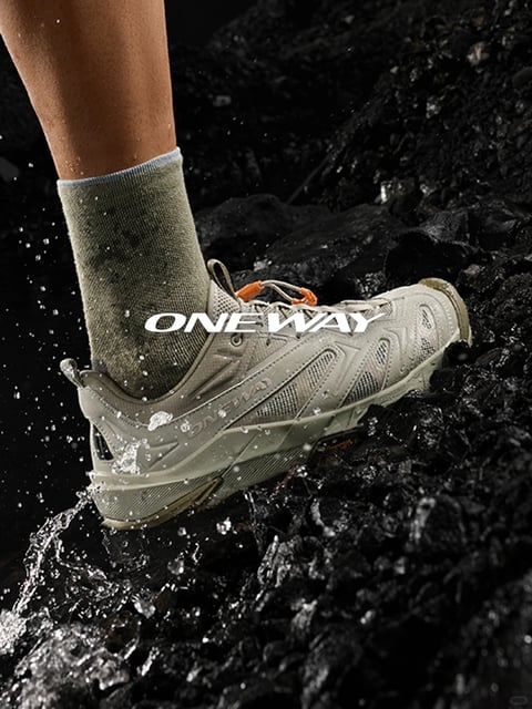 The image presents a dynamic close-up of a person's lower leg and foot stepping into shallow, splashing water over dark, wet volcanic rocks, capturing a rugged outdoor action scene. The composition is tightly framed on the footwear and ankle, with a shallow depth of field that sharply focuses on the shoe and splashing water while blurring the rocky background, creating a sense of immediacy and immersion. Visually, the scene is rendered in a high-contrast, photorealistic style with a dominant palette of muted olive greens from the sock and shoe, deep charcoal blacks from the wet rocks, and crisp white highlights from the water spray and text. Lighting appears directional from above and slightly left, creating hard, glossy reflections on the wet rock surfaces and casting sharp shadows within the shoe's mesh textures, emphasizing its technical construction. The materials are vividly rendered: the sock shows a ribbed, absorbent knit texture, the shoe combines durable synthetic overlays, breathable mesh panels, and a rugged outsole, all glistening with water droplets that convey a tactile sense of wetness and performance. The text 'ONE WAY' is prominently displayed in bold, white, sans-serif capital letters overlaid across the shoe's midsection, acting as a brand or model identifier integrated into the action. The overall mood is energetic and adventurous, with the water splash adding kinetic energy and the tight framing focusing attention on the product's durability in an outdoor, elemental context.
