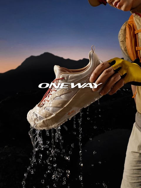 The image presents a photorealistic, high-contrast outdoor adventure scene featuring a close-up of a person holding a single light beige hiking shoe. The shoe is dripping with water, with droplets suspended mid-air, emphasizing its waterproof quality against a backdrop of dark, silhouetted mountains under a twilight sky transitioning from deep blue to warm orange near the horizon. The composition is tightly framed on the shoe and the person's gloved hand, which wears a yellow fingerless glove, creating a strong focal point. The lighting is dramatic and directional, likely from a low-angle source mimicking the setting sun, casting hard highlights on the wet shoe surface and the person's skin while plunging the background into deep shadow. The color language is dominated by earthy neutrals—the shoe's beige, the dark rocky background—with accents of warm orange in the sky and glove, and cool blue in the upper atmosphere, creating a high-contrast, moody palette. Text reading 'ONE WAY' in bold, white, sans-serif capital letters is overlaid centrally across the shoe, serving as a brand identifier. The texture rendering is highly detailed, showcasing the shoe's mesh and synthetic overlays, the slick wetness of the water droplets, and the fabric of the glove. The overall mood is rugged, resilient, and aspirational, conveying a sense of performance and durability in harsh natural environments.