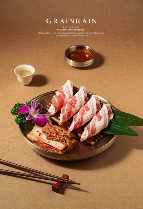 The image presents a meticulously styled food photograph centered on a metallic platter holding four slices of raw, marbled beef arranged in a fan shape on a wooden slat grid, accompanied by a portion of kimchi, a purple orchid, and green leaves for garnish. To the upper left, a small white teacup contains a light amber liquid, while a metallic dish with a dark reddish-brown dipping sauce sits above the main platter. In the bottom left foreground, a pair of dark wooden chopsticks rests on a small wooden holder. The composition is set against a textured, light brown paper-like background, with a clear, readable English text block at the top center: 'GRAINRAIN', followed by smaller text 'PHOTOGRAPHY' and 'KNOWLEDGE AND FOOD CULTURE', and a paragraph below stating 'Delicious food is for enjoyment. Watching, smelling and even thinking can be regarded as a kind of enjoyment.' The visual aesthetic is highly photorealistic with a professional, commercial food photography style, employing a warm color palette dominated by earthy browns, the pink and white of the beef, and accents of purple and green. Lighting is soft and directional from the upper left, casting gentle shadows that enhance the three-dimensional form of the objects and highlight the glistening texture of the meat and sauces. Textures are rendered with high detail, from the marbling on the beef and the fibrous kimchi to the matte ceramic of the cup and the grain of the paper background. The composition uses a balanced, slightly overhead arrangement with a shallow depth of field that keeps the main platter in sharp focus while subtly softening the background elements. Shape language combines the organic forms of the food and flower with the geometric lines of the platter, grid, and chopsticks. The overall mood is elegant, appetizing, and contemplative, evoking a sense of refined culinary appreciation.