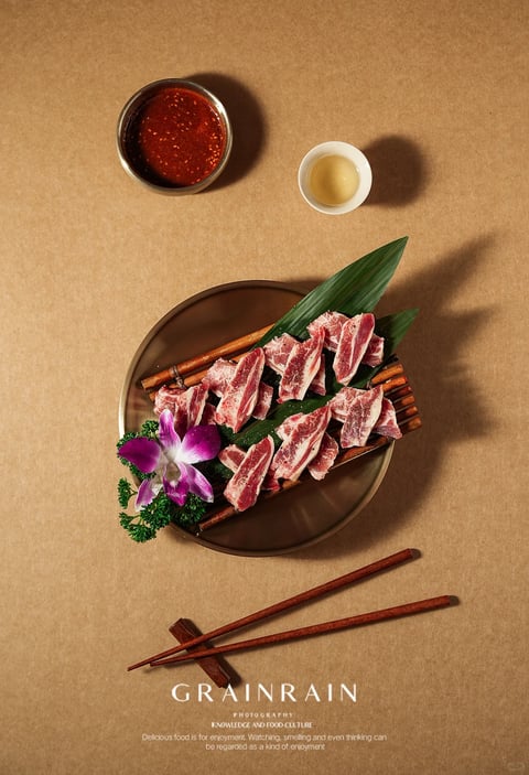 This is a high-angle, top-down still life photograph of a Japanese-style barbecue meat dish presented on a smooth, dark brown circular plate against a textured, light tan craft paper background. The main subject is a pile of raw, marbled beef slices (likely wagyu or ribeye) arranged on a small bamboo raft, accented with a single purple-and-white orchid, green parsley, and a large green leaf. To the upper left, a small, dark bowl contains a glossy, red chili dipping sauce with sesame seeds, and to the upper right, a small white ceramic cup holds a pale yellow liquid, possibly sake or tea. At the bottom, a pair of dark wooden chopsticks rests on a chopstick holder. Visible text appears at the bottom center in white serif font: the brand name "GRAINRAIN," followed by "PHOTOGRAPHY" in smaller letters, and a three-line caption below reading "KNOWLEDGE AND FOOD CULTURE / Delicious food is for enjoyment. Watching, smelling and even thinking can / be regarded as a kind of enjoyment." The visual aesthetic is photorealistic with a professional food photography style, featuring soft, directional lighting from the upper left that casts defined, elongated shadows to the right, creating a warm, inviting mood. The color palette is earthy and warm, dominated by the tan background, deep browns of the plate and chopsticks, the rich reds and pinks of the meat, and pops of green and purple from the garnish. Textures are highly detailed, from the fibrous grain of the paper background and bamboo to the moist, fatty marbling of the meat and the glossy surface of the sauce. The composition is balanced and symmetrical, using negative space to highlight the dish, with a shallow depth of field that keeps the entire setup in sharp focus. The shape language contrasts organic forms (meat slices, flower petals, leaves) with geometric elements (circular plates, straight chopsticks), and the overall atmosphere is elegant, appetizing, and culturally refined.