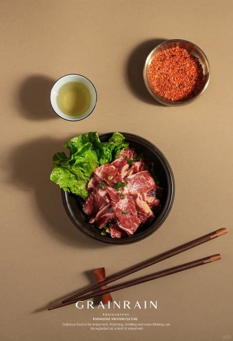 This is a top-down, photorealistic studio photograph of a food preparation scene, composed with deliberate minimalism and strong geometric balance. The main subject is a dark, matte ceramic bowl centered in the lower half, containing raw, marbled slices of red meat (likely beef) resting on a bed of vibrant green lettuce leaves and garnished with finely chopped green onions. Above the bowl, to the left, is a small, white ceramic bowl with a thin dark blue rim, holding a clear, pale yellow liquid, possibly oil or broth. To the right, a clear glass bowl contains a coarse, bright red spice blend, likely chili flakes. In the lower foreground, a pair of dark brown wooden chopsticks rests diagonally on a simple wooden chopstick holder. The background is a flat, uniform, warm beige surface. At the bottom, white text in a clean, sans-serif font reads "GRAINRAIN", below which is smaller text: "PHOTOGRAPHY", "KNOWLEDGE AND FOOD CULTURE", and the English phrase: "Delicious food is for enjoyment. Watching, smelling and even thinking can be regarded as a kind of enjoyment." The lighting is soft and directional from the top left, casting soft but defined shadows that give the objects depth and a grounded feel, creating a warm, inviting, and contemplative mood focused on the raw, natural textures of the ingredients. The composition uses negative space effectively, with the elements arranged in a balanced, triangular layout that guides the eye through the components of a potential meal.