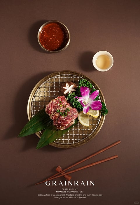 The image presents a meticulously styled food photography scene with a top-down, flat-lay composition centered on a round, dark-rimmed plate. The plate features a metal wire grill, upon which rests a rectangular slab of raw, marbled red meat garnished with finely chopped green herbs, placed atop two large, vibrant green leaves. To the side of the meat, a small pile of dark green curly parsley, a halved shiitake mushroom with a decorative star cut, a slice of lemon, and a vibrant pink and white orchid flower create an elegant garnish. Above the plate, two small bowls are arranged: a dark bowl containing a textured, reddish-brown dipping sauce with visible seeds or grains, and a small, white bowl holding a clear, light-amber liquid. Below the plate, a pair of dark wooden chopsticks rests diagonally on a simple wooden chopstick rest. The background is a solid, deep brown with a subtle matte texture. The lighting is soft and directional, casting gentle shadows that give depth to the food and bowls, enhancing the rich, warm color palette of deep browns, reds, and greens, contrasted by the bright pink of the orchid and the white of the small bowl. Visible text at the bottom reads "GRAINRAIN" in large, serif, all-caps white font, followed by smaller text: "PHOTOGRAPHY", "KNOWLEDGE AND FOOD CULTURE", and the phrase "Delicious food is for enjoyment. Watching, smelling and even thinking can be regarded as a kind of enjoyment" in a smaller, white sans-serif font, serving as a branding and thematic caption. The overall aesthetic is polished, photorealistic, and sophisticated, with a focus on texture—from the grain of the meat and wood to the gloss of the sauces—and a balanced, harmonious arrangement that conveys a mood of refined culinary appreciation.