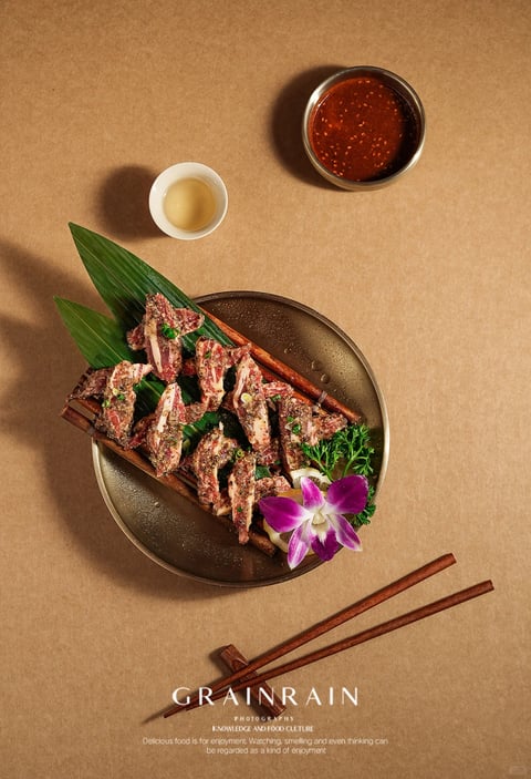 This is a high-angle, top-down food photography shot centered on a plated dish of marinated, uncooked lamb chops presented on a bamboo mat over large green leaves, arranged on a dark, textured ceramic plate. The plate is accented with a sprig of green herbs and a purple orchid flower. Above the plate to the left sits a small white cup with a pale yellow liquid, and to the upper right, a small dark bowl filled with a rich, reddish-brown dipping sauce speckled with seeds. Below the plate, a pair of wooden chopsticks rests on a chopstick holder. The text 'GRAINRAIN' in large, white, serif font is overlaid near the bottom, with smaller text below reading 'PHOTOGRAPHY', 'KNOWLEDGE AND FOOD CULTURE', and a descriptive sentence. The overall aesthetic is photorealistic with a warm, inviting color palette dominated by earthy browns, deep reds, and vibrant greens, enhanced by soft, directional lighting from the top left that creates gentle shadows and highlights the textures of the food and materials. The composition is balanced and clean, with a shallow depth of field focusing sharply on the central dish, set against a uniform, light brown, textured background.