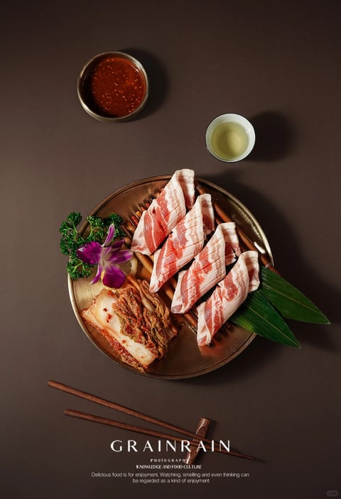 The image presents a photorealistic, top-down, overhead composition of a culinary arrangement centered on a dark bronze, textured round tray, featuring thick, marbled raw pork belly slices with scored diamond patterns, reddish kimchi, and decorative elements like a purple orchid and green parsley. Accompanying the main tray are a small, rustic ceramic bowl of reddish-brown chili dipping sauce and a smaller white ceramic cup of pale yellow liquid, likely a light broth or tea. The scene is set against a dark, matte brown surface with subtle texture. Visible text in the lower portion includes 'GRAINRAIN' in elegant, serif, uppercase white letters, below which 'PHOTOGRAPHY' appears in smaller sans-serif, followed by 'KNOWLEDGE AND FOOD CULTURE', and a descriptive phrase: 'Delicious food is for enjoyment. Watching, smelling and even thinking can be regarded as a kind of enjoyment,' all in white, creating a clean, editorial layout. The lighting is directional from the upper left, casting soft, defined shadows that enhance the three-dimensionality and rich textures of the food and tableware, conveying a sophisticated, high-end dining atmosphere with warm color temperature and high contrast.