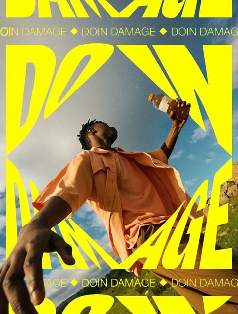 The image presents a low-angle, full-shot photograph of a Black man with short dreadlocks, wearing a loose, short-sleeved peach button-down shirt and dark pants, captured against a bright blue sky with soft white clouds and a glimpse of green, rocky terrain at the bottom right. His body is angled away from the camera, with his head tilted back looking upward, his right arm extended down towards the viewer in the foreground with an open palm, and his left arm raised holding a clear plastic bottle of iced coffee with an orange cap. The composition is dominated by large, bold, sans-serif typography in a vibrant, high-saturation yellow that frames and overlaps the subject; the words 'DOIN DAMAGE' are prominently displayed in a fragmented, distorted arrangement across the image, with smaller, repeating horizontal banners of the same text in the upper and lower thirds. The visual aesthetic is a blend of editorial photography and graphic design, characterized by high contrast, sharp details on the subject and text, and a slightly grainy, film-like texture. The lighting is bright, direct sunlight, creating strong highlights on the shirt and skin, and deep shadows, particularly on the subject's face and under the extended arm, contributing to a dynamic and high-energy mood. The shape language contrasts the organic, fluid forms of the subject's pose and clothing with the hard, geometric, and deliberately warped edges of the yellow text overlays. The overall atmosphere is confident, bold, and contemporary, with a sense of upward momentum and visual impact.