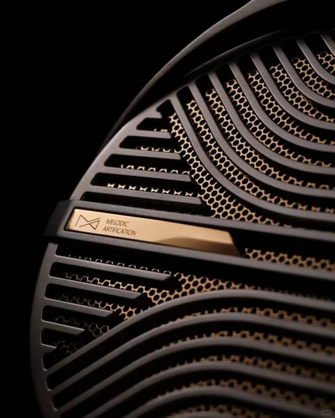 The image presents a close-up, angled view of a sophisticated audio speaker grille, showcasing a luxurious industrial design aesthetic. The main subject is a curved, dark gray or black outer grille with undulating, parallel slats that create a sense of dynamic movement and depth. Behind this, a secondary, finely perforated golden honeycomb mesh is visible, adding a layer of intricate texture. The composition is tightly cropped, emphasizing the interplay of forms and materials against a stark, deep black background. A prominent, rectangular, brushed gold-colored metal plate is affixed horizontally to the grille. It features a geometric logo resembling a stylized 'M' or an abstract audio wave on the left, followed by the clearly readable text 'MELODIC ARTIFICATION' in a clean, sans-serif, uppercase font, which serves as a brand or model identifier, reinforcing the product's artistic and musical theme. The visual aesthetic is highly photorealistic, with meticulous attention to material rendering. The color language is defined by a high-contrast palette of deep, neutral blacks and dark grays against warm, reflective gold tones, creating a feeling of premium quality. Lighting appears to come from the upper left, casting soft, directional highlights along the edges of the dark slats and the gold plate, while creating deep, soft shadows within the recesses, enhancing the three-dimensional form and tactile quality. The texture is a study in contrast: the smooth, matte finish of the dark outer grille, the precise, geometric pattern of the porous honeycomb mesh, and the subtle, linear brushed texture of the metallic plate. The composition uses a shallow depth of field, with the gold plate and its text in sharp focus while the grille patterns recede into a softer focus, guiding the viewer's eye. The shape language combines the organic, wave-like curves of the main grille with the strict, geometric repetition of the honeycomb pattern and the sharp lines of the metal plate. The overall mood is one of sophisticated, elegant minimalism with a strong sense of engineered luxury and focused craftsmanship.