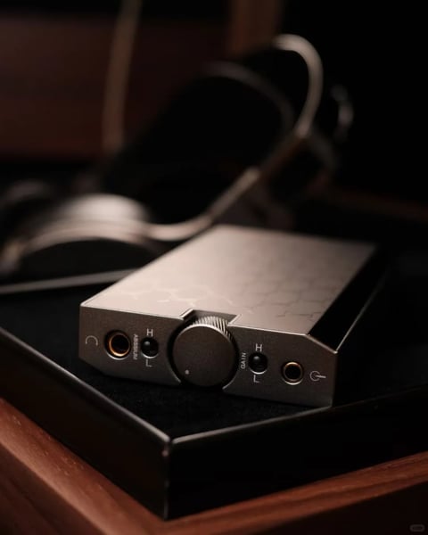 The image presents a close-up, photorealistic still life of a premium portable audio device, likely a headphone amplifier or DAC, positioned centrally on a dark, textured surface within a shallow black tray. The device is rendered in a matte silver metal with a subtle honeycomb pattern across its top surface, featuring a prominent central volume knob with fine knurling. Its front face includes two gold-ringed audio jacks, toggle switches labeled 'ABSSUM' with 'H' and 'L' settings on the left and 'GAIN' with 'H' and 'L' settings on the right, and symbolic icons resembling a power symbol and a circular arrow. In the softly blurred background, a pair of over-ear headphones with a silver headband and dark earcups rests on what appears to be a wooden surface, creating a shallow depth of field that emphasizes the main device. The lighting is directional from the upper left, casting soft, defined highlights along the device's edges and the volume knob's ridges while leaving the right side in deep, warm shadow, creating high contrast with a predominantly monochromatic palette of silvers, blacks, and dark browns, accented only by the gold of the audio jacks. The overall composition is tightly framed with a slight low-angle perspective, emphasizing the device's geometric, angular form and tactile metallic textures, evoking a mood of sophisticated, high-fidelity audio craftsmanship.