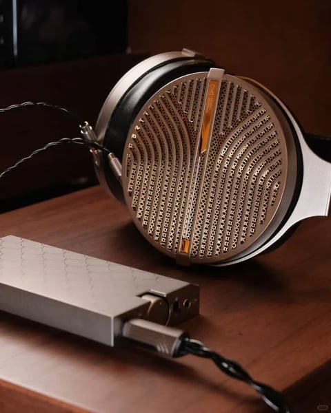 This is a photorealistic, close-up studio-style image with a shallow depth of field, focusing on a high-end open-back headphone and a portable DAC/amp on a dark wooden surface. The main subject is the right earcup of the headphone, angled towards the viewer, showcasing its intricate, wave-patterned metal grille with a honeycomb mesh underneath and a vertical amber-gold accent strip bearing a small hourglass logo. The headphone's frame is brushed silver metal with a black leather ear pad visible. A braided black audio cable connects to the earcup via two silver plugs. In the foreground, slightly out of focus, lies a rectangular, silver portable DAC/amp with a textured top surface and a USB-C cable plugged into its side. The composition is arranged diagonally, with the headphone dominating the upper right and the DAC extending from the lower left towards the center, creating a sense of depth. The lighting is soft and directional, coming from the upper left, casting gentle highlights on the metallic surfaces and creating warm amber reflections within the grille's recesses, while the background falls into deep, soft-focus shadow. The color palette is sophisticated and muted, dominated by cool silver and warm bronze-gold tones against the rich, dark brown of the wooden table, with high contrast between the illuminated metallic objects and the shadowy environment. There is no obvious readable text beyond the small logo on the amber strip. The overall mood is luxurious, technical, and serene, emphasizing the craftsmanship and material quality of the audio equipment.