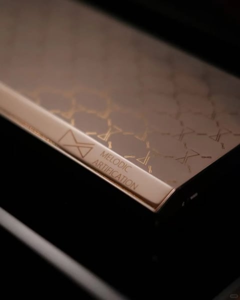 The image presents a close-up, high-angle view of a rectangular object, likely a luxury product box or a sophisticated electronic device casing, set against a deep, dark background that creates a dramatic, low-key atmosphere. The primary visual element is the object's top surface and angled edge, which features a complex, repeating pattern of thin, interconnected golden geometric shapes—resembling abstract line-art molecules or stylized runes—etched onto a soft, matte, dusky rose or taupe surface. The object's side edge is defined by a polished, reflective rose-gold metallic trim that catches the light. This trim bears the engraved text "MELODIC ARTIFICATION" in a clean, modern, all-caps sans-serif font, accompanied by a small, minimalist geometric logo of two interlocking triangles. The text is precisely placed and integrated into the metallic band, serving as a brand or product identifier. The focus is critically sharp on the text and the immediate edge detail, with the patterned surface and background falling into a soft, shallow depth of field. The lighting is directional and soft, coming from the upper left, creating a warm, luminous highlight along the rose-gold edge and casting subtle gradients across the matte surface, enhancing its tactile, velvety impression. The overall aesthetic is one of minimalist luxury, combining geometric precision with warm, muted tones and a high-contrast, moody presentation.