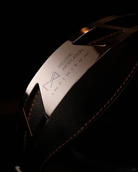 The image presents a close-up, low-angle shot of a high-end audio headphone's ear cup against a deep, near-black background, emphasizing a premium, luxurious aesthetic. The main subject is a curved, metallic band featuring a polished, rose-gold or champagne-gold finish that catches the light, contrasting with the adjacent ear cup wrapped in textured black leather with visible, precise orange or gold stitching along its edge. The composition is tightly framed, focusing on the interplay of materials and the engraved text. The visible text is rendered in a clean, sans-serif font and reads 'MELODIC ARTIFICATION' stacked above 'INFINITUM', with a stylized 'M' logo to the left; the text is etched into the metallic surface, appearing darker than the metal and serving as a brand or product identifier. The focus is sharply on this metallic band and its engravings, with the leather texture and stitching slightly softer but still distinct. The visual aesthetic is photorealistic with a sleek, product-photography rendering style, highlighting material quality. The color language is dominated by deep blacks and shadows, punctuated by the warm, reflective metallic tones and the subtle orange accents of the stitching, creating a high-contrast, dramatic palette. The lighting is directional and soft, likely from an upper-left source, casting gentle highlights on the metal's curvature and creating deep shadows that enhance the three-dimensional form and luxurious mood. The texture and material feel are highly detailed, contrasting the smooth, cool polish of the metal with the fine grain and tactile warmth of the leather. The composition uses a diagonal arrangement and shallow depth of field to guide the eye to the branded metal element. The shape language is a mix of the organic curve of the headphone and the sharp, geometric lines of the engraved logo and text. The overall mood is sophisticated, exclusive, and moody, conveying a sense of high-fidelity craftsmanship and premium audio experience.