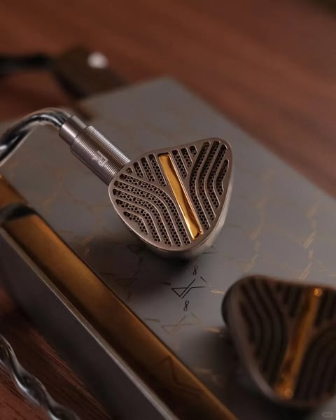 The image features a pair of high-end in-ear monitors (IEMs) as the main subject, presented in a close-up, shallow depth-of-field composition. One IEM is in sharp focus, resting diagonally atop a metallic, possibly silver or gunmetal-colored device with a brushed finish and subtle engraved patterns, while the second IEM is softly blurred in the lower right foreground. The primary IEM showcases a distinctive, triangular-shaped faceplate with a complex, open-back grille design. This grille consists of concentric, wave-like slats radiating from a central, vertical gold-accented bar, all rendered in a matte silver or titanium finish with precise, machine-cut detailing. A braided, translucent silver cable connects to the IEM via a metallic connector with a ribbed texture. The lighting is soft and directional, likely from the upper left, casting gentle highlights on the metallic surfaces and creating deep, soft shadows that accentuate the three-dimensional form and intricate textures. The color palette is dominated by cool metallic tones—silver, gunmetal, and chrome—contrasted sharply by the warm, polished gold accent on the IEM's faceplate. The material feel is highly tactile, emphasizing precision-engineered metal with a mix of matte, brushed, and polished finishes. Compositionally, the sharp focus on the central IEM's intricate grille against the blurred foreground and background creates a strong sense of depth and draws the eye to the product's detailed craftsmanship. The overall mood is luxurious, technical, and sophisticated, conveying a sense of premium audio engineering and aesthetic refinement.