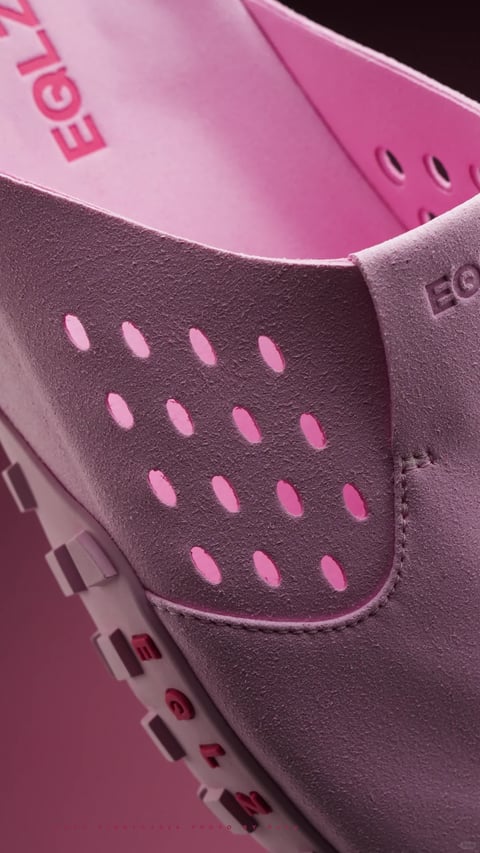 This is a close-up, high-angle photographic shot focusing on the heel and upper section of a pink suede shoe, with a shallow depth of field that emphasizes the tactile materials and construction details. The primary subject is the textured, mauve-pink suede upper, which features a precise pattern of oval perforations revealing a brighter, saturated pink inner lining. The composition is tightly framed, with the shoe's structure creating strong diagonal lines and curves, and the background consists of a solid, matching dark mauve surface that creates a monochromatic and cohesive color field. Visible text includes partial embossed lettering on the inner side of the shoe's tongue, where the red letters 'EGI' are clearly readable, suggesting a brand name or model identifier, and the letters 'EQ' are debossed into the suede on the outer side of the heel counter. The image exhibits a photorealistic aesthetic with a soft, diffused lighting approach coming from the upper left, casting gentle shadows that accentuate the suede's nap and the three-dimensional quality of the perforations and stitching. The color language is dominated by a sophisticated, muted mauve-pink palette with a pop of vibrant fuchsia-pink from the lining, creating a tonal contrast with high saturation within a narrow hue range. The material feel is highly tactile, emphasizing the soft, fibrous texture of the suede against the smoother, perforated inner layer and the rigid, molded detail of the sole edge visible in the lower left corner. The mood is elegant, focused, and subtly luxurious, conveyed through the precise craftsmanship, soft lighting, and tonal color harmony.