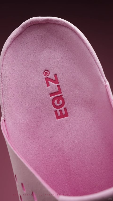 This image is a photorealistic, close-up studio shot focusing on the insole of a light pink shoe, likely a sneaker, presented against a dark, muted burgundy or plum background. The composition is tightly framed, angled from above, and dominated by the soft, textured surface of the insole, which fills most of the frame. The visual aesthetic is clean, modern, and minimalist, with a soft, diffused light coming from the upper left, casting gentle, subtle shadows that enhance the material's tactile quality. The color language is monochromatic and warm, centered on a soft, desaturated pink with a fine, suede-like or microfiber texture, creating a plush, velvety impression. The dominant color is this soft pink, with contrast provided by the darker, recessed logo text and the deep, warm-toned background. The text, which reads 'E·G·N·I' in a stylized, vertical arrangement with a registered trademark symbol (®) beside it, is debossed or printed in a darker shade of pink or red directly onto the insole's surface, serving as a brand identifier. The focus is sharp on the central text and the immediate surrounding texture of the insole, with the edges of the shoe and the background softly blurred, creating a shallow depth of field that emphasizes the material detail and branding. The overall shape language is organic, following the curved contour of the shoe's interior, with soft edges and a smooth, continuous surface. The mood is calm, sophisticated, and subtly luxurious, evoking a sense of comfort and refined craftsmanship.