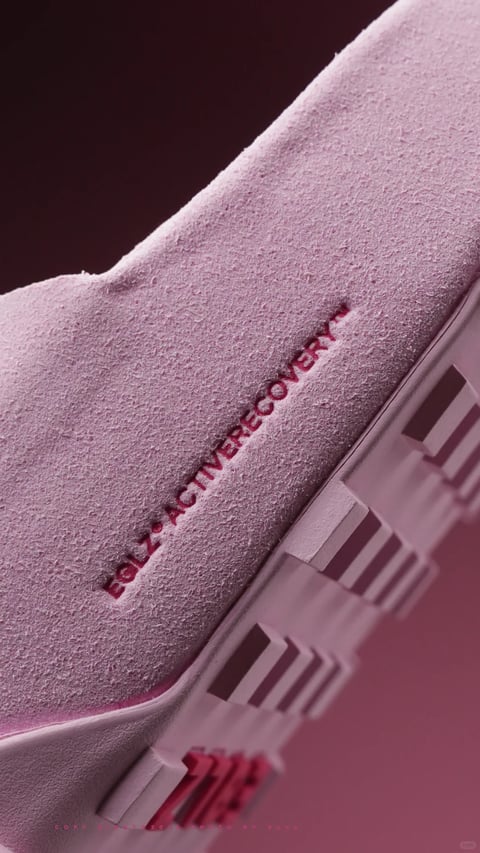 The image is a close-up, angled studio photograph of a light pink, textured sneaker, likely a recovery slide, captured against a dark, contrasting background. The composition is dynamic, with the shoe's upper and sole filling the frame diagonally, creating a sense of depth through a shallow depth of field that blurs the background and foreground elements. The primary visual element is the shoe's material, which appears to be a soft, granular foam or suede-like texture, rendered with fine detail that emphasizes its tactile, cushioned quality. Visible text is debossed into the side of the shoe in a darker magenta hue, reading "EDLZ ACTIVERECOVERY" in a clean, sans-serif font; this text acts as a branding element and emphasizes the product's functional purpose. The lighting is soft and directional, coming from the upper left, which creates subtle shadows that enhance the texture and form of the shoe, conveying a mood of calm, comfort, and premium quality. The color palette is monochromatic, dominated by soft pinks and mauves with deep, muted magenta accents, characterized by low saturation and a gentle, warm temperature. The shape language combines the organic, curved form of the shoe's upper with the geometric, blocky patterns of the sole, featuring sharp edges and defined lines. The overall visual aesthetic is a blend of photorealistic product photography with a soft, almost painterly focus on texture and light, creating a sophisticated and serene atmosphere.