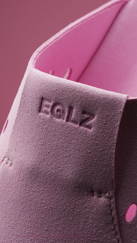 This is a close-up, photorealistic image focusing on the corner of a pink leather bag. The composition is tight, framing the bag's flap and a portion of its body against a matching pink background, creating a monochromatic and cohesive visual. The dominant color is a vibrant, saturated magenta-pink, with subtle tonal variations that define the bag's form through soft, directional lighting from the upper left. This lighting casts gentle shadows, enhancing the tactile impression of the materials. The bag features two distinct textures: the main body has a smooth, slightly glossy leather finish, while the overlapping flap is made of a contrasting, napped suede material with a visible, fuzzy texture. The edges are neatly finished, with precise stitching visible along the seams. Embossed deeply into the suede flap is the text 'EGLZ', rendered in a bold, geometric, sans-serif typeface. The letters are debossed, creating a recessed effect that plays with the light and shadow. The overall mood is modern, stylish, and focused on material quality and detail.