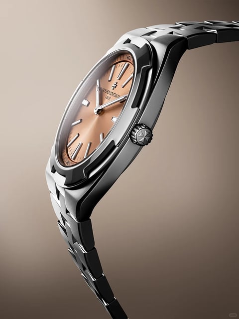The image presents a studio-lit, close-up shot of a luxury wristwatch, captured in a dynamic, floating profile view against a soft, gradient beige-to-brown background. The main subject is a Vacheron Constantin watch, angled to showcase its side profile, crown, and dial. The form features a polished, silver-toned stainless steel case with a distinctive, integrated bracelet design where the links flow seamlessly from the lugs, creating a unified, curved silhouette. The materials exhibit high-quality finishing, with the case and bracelet displaying a mix of brushed and polished surfaces that catch the light, highlighting the geometric, angular design of the links. The dial is a warm, salmon or copper color with a sunburst finish, marked by silver-toned, applied baton hour markers and hands. Visible text on the dial includes 'VACHERON CONSTANTIN' and 'GENEVE' below the brand's Maltese cross logo at the 12 o'clock position, and small numerals '120' and '160' on the flange near the 7 and 11 o'clock positions, respectively. The crown is prominently featured, with a textured grip and the Maltese cross emblem engraved on its end. Construction details reveal precise machining, with clean lines, a bezel that integrates smoothly into the case, and a crown guard that protects the winding mechanism. The overall style is realistic and modern luxury, emphasizing technical precision and elegant design, suitable for a high-end fashion or horological context.
