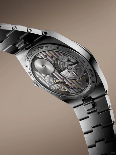 This image presents a high-angle, close-up view of the caseback of a luxury wristwatch, set against a smooth, neutral gradient background transitioning from warm taupe to cool gray. The main subject is the intricate mechanical movement visible through a sapphire crystal caseback, framed by a brushed and polished stainless steel case and integrated bracelet. The composition is diagonal, emphasizing the watch's curved form and the depth of the movement. Visible text is engraved on the steel caseback perimeter and on components within the movement. On the steel rim, in a clean, sans-serif font, reads 'VACHERON CONSTANTIN' at the top, '2500V' and 'P950' on the right side, '345T952' further down, 'Limited Edition No 01/250' along the bottom curve, 'SWISS MADE' and '50M' on the left side. Inside the movement, smaller gold-filled text on the bridges reads 'VACHERON CONSTANTIN', 'GENEVE', and 'TWENTY-SIX (26) JEWELS'. The focus is sharpest on the mechanical movement, showcasing its exquisite construction with a large, silver-toned, radially brushed rotor bearing a Maltese cross motif, visible gear trains, ruby jewels set in polished sinks, and bridges with fine, linear Geneva stripe finishing. The form is a sophisticated, integrated sports watch silhouette with flowing lugs that seamlessly connect to the multi-link bracelet, which features a combination of brushed surfaces and polished chamfered edges. The materials are primarily stainless steel with a high-quality finish, sapphire crystal, and internal components of brass, steel, and synthetic ruby. Functional cues are evident in the self-winding rotor mechanism and the screw-fastened caseback. The style is hyper-realistic and detailed, conveying a sense of precision engineering and luxury craftsmanship, with context signals suggesting a high-end, collectible timepiece.