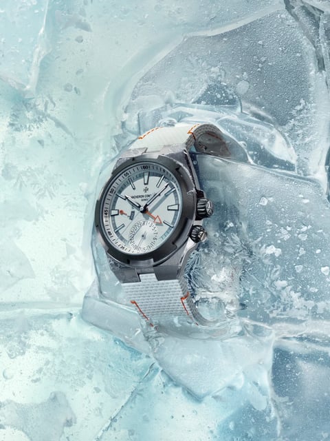 A high-end Vacheron Constantin Overseas Dual Time wristwatch is dramatically embedded in a block of translucent, fractured ice. The watch is positioned at a slight angle, with its silver-toned, brushed steel case and integrated bracelet partially submerged in the frozen medium. The ice features intricate internal patterns of air bubbles and feathery frost crystals, with cool blue and white tones dominating the scene. The watch face is white with applied, luminescent baton hour markers and a silver chapter ring with minute numerals. It displays two time zones via stacked hour and minute hands in dark metal, with a small orange-tipped GMT hand. A date subdial is located at 6 o'clock, and a power reserve indicator sits at 9 o'clock. The bezel has a distinctive six-sided design with polished bevels. The bracelet is a textured, light gray or off-white fabric strap with visible orange contrast stitching near the lugs. Two crowns are visible on the right side of the case: one for main adjustments and a secondary pusher for the dual time function. Visible text on the dial includes the brand name 'VACHERON CONSTANTIN GENEVE' below the 12 o'clock marker and the Swiss cross emblem at 12 o'clock. Numerical markers are present on the inner bezel ring (05, 10, ..., 60) and on the date subdial (1 through 31). The focus is sharp on the watch dial and bezel, with the ice texture becoming more abstract and blurred in the foreground and background, emphasizing the watch as the central subject within its extreme, cold environment.