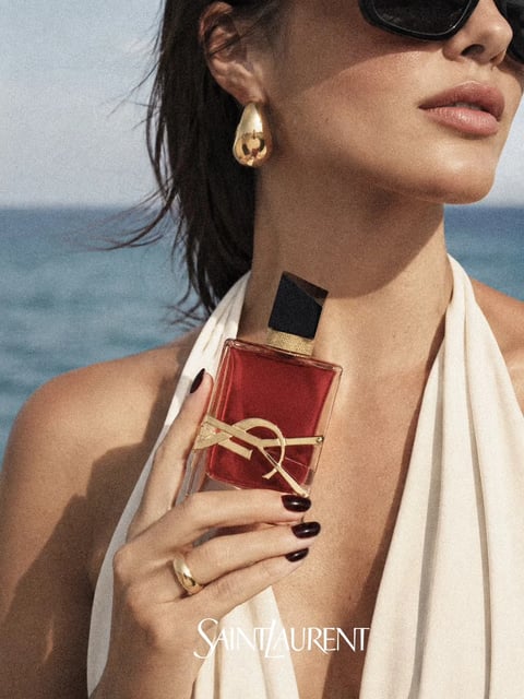 A cinematic close-up photograph features a woman with dark, windswept hair, positioned against a backdrop of the calm blue ocean and a bright, hazy sky. The composition focuses on her upper torso and hand, which elegantly holds a rectangular perfume bottle filled with a vibrant red liquid, positioned near her neck. Visible text at the bottom of the image reads 'SAINT LAURENT' in a classic, white serif font, acting as the brand identifier. The perfume bottle has a distinct geometric form: a clear glass rectangle with a sharp, black, asymmetrical cap. A prominent, sculptural gold YSL monogram wraps around the middle of the bottle, creating a tactile and metallic contrast against the smooth glass. The woman’s hand, with dark-polished nails, showcases a polished gold ring. Her attire consists of a white, draped halter-neck top with soft folds, and she wears a large, organic-shaped gold teardrop earring. The overall style is luxurious and realistic, with warm, natural lighting that highlights the textures of the skin, the glossy surfaces of the gold accessories, and the reflective glass of the bottle, while the ocean background provides a serene, summery context.