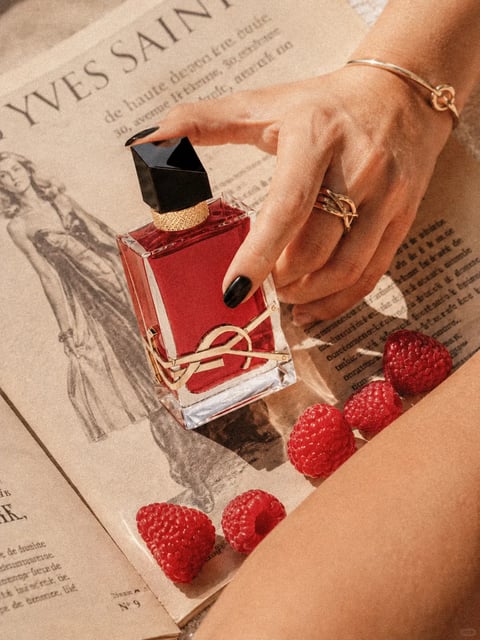 The image features a luxurious red perfume bottle held by a hand adorned with a gold ring and bracelet, set against a backdrop of vintage-style newspapers. The perfume bottle, with its rectangular glass body and black geometric cap, is the central focus, its deep red liquid visible through the glass. The hand, with black-painted nails, grips the bottle, adding a sense of interaction and elegance. The newspapers in the background, with visible text including 'YVES SAINT' and other French phrases, contribute to a sophisticated, classic ambiance. The scene is complemented by scattered red raspberries, adding a vibrant, fresh contrast to the warm tones of the bottle and skin. The overall composition is rich in texture, from the smooth glass of the bottle to the rough paper of the newspapers and the soft, juicy appearance of the raspberries, creating a visually appealing and indulgent mood.