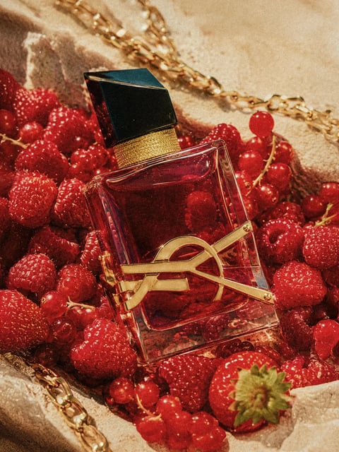 A high-angle, close-up shot features a rectangular glass perfume bottle filled with a deep, ruby-red liquid, lying diagonally amidst a lush, textured bed of fresh red berries including raspberries, strawberries, and currants, set against a backdrop of rough, light-beige sand or stone. The bottle, the primary focal point, has a glossy black rectangular cap atop a textured gold collar, and its front is adorned with a prominent, raised gold metal emblem resembling an intertwined 'Y' and 'C' (or a stylized 'YSL' monolith), which catches the light. The composition is rich and indulgent, with the warm, saturated reds of the fruit and perfume contrasting sharply with the neutral background and metallic accents. A delicate, gold chain link necklace snakes through the scene in the upper right and lower left, adding a touch of ornate, luxurious detail. There is no visible text or branding on the bottle or within the scene. The focus is sharpest on the perfume bottle's gold emblem and the surrounding berries, with a shallow depth of field that softly blurs the background elements, emphasizing the tactile textures of the glossy glass, juicy fruit, and granular sand. The overall style is luxurious, realistic, and opulent, with a warm color palette dominated by deep reds, golds, and creamy beiges, suggesting an indulgent or romantic context.