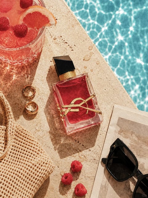 A sun-drenched poolside flat-lay composition featuring a bottle of YSL Libre Eau de Parfum as the central subject, resting on a textured beige stone surface beside shimmering turquoise pool water. The perfume bottle showcases a rectangular clear glass form with faceted edges containing a warm rose-pink liquid, topped with a geometric black asymmetrical cap and accented with gold hardware including a textured neck band and the iconic interlocking YSL logo wrapped around the bottle in polished gold metal. Surrounding lifestyle accessories include two gold rings with minimal geometric settings, a cream crocheted woven bag with open-mesh texture in the lower left, a cut crystal glass filled with pink grapefruit cocktail garnished with fresh raspberries and a citrus wheel in the upper left corner, and black acetate sunglasses resting on a magazine in the lower right. Three fresh raspberries are scattered near the bottom of the frame, echoing the cocktail garnish. Strong directional sunlight casts defined shadows across the stone surface, creating bright highlights on the glass bottle and metallic gold elements while the pool water displays dappled light refraction patterns. The overall aesthetic is luxurious summer leisure with warm golden tones contrasting against cool aqua water, rendered in a realistic photographic style with rich textural detail across the stone, crochet, and glass surfaces. There is no prominent readable text visible in the composition beyond the recognizable YSL brand logo integrated into the perfume bottle design.