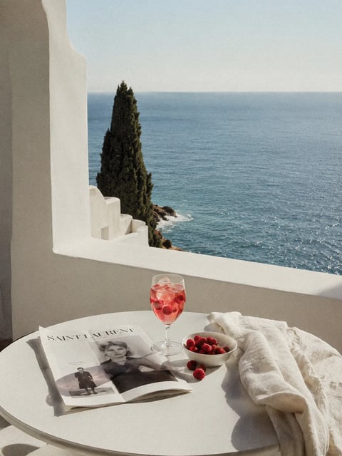 A serene, sunlit coastal scene viewed from a white architectural terrace, where a round white table in the foreground holds a glass of pink iced beverage with raspberries, a small white bowl filled with fresh raspberries, and an open Saint Laurent magazine. The background reveals a calm, deep blue sea meeting a clear sky, framed by a white stucco wall with geometric steps and a tall, dark green cypress tree. Visible text on the open magazine includes the brand name 'SAINT LAURENT' in bold, uppercase letters at the top of the left page, with additional smaller, illegible text below, establishing a luxury fashion context. The overall composition balances minimalist, clean lines of the white architecture with organic textures of the tree and fruit, creating a tranquil, high-end summer aesthetic.