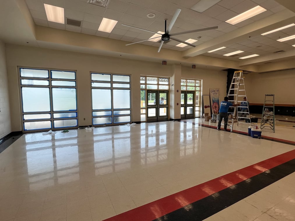This is an image from Halls Quality Painting company, located in Fernandina Beach, FL. The image is an interior of a commercial building with freshly cleaned and polished floors, large glass windows covered for painting, and workers on ladders preparing walls for painting at Hall’s Quality Painting project site.