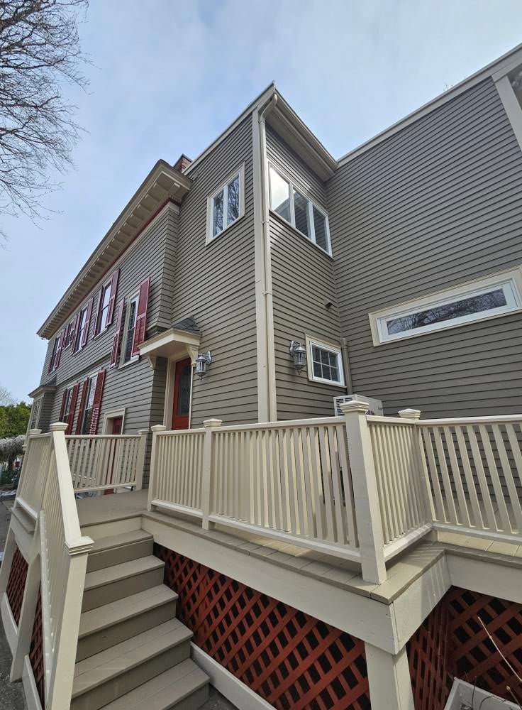 This is an image from WC Painting Inc, located in Massachusetts. The photo shows a two-story home exterior with beige siding, red shutters, and cream-colored trim and railings freshly painted to highlight the entryway and deck area.