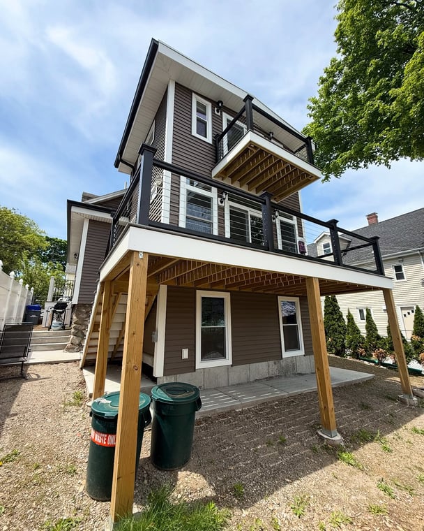 This is an image from EVM Home Improvement & Painting, located in Massachusetts. The photo shows a two-story home with newly painted dark brown siding, white trim, and a modern elevated deck with black railing.