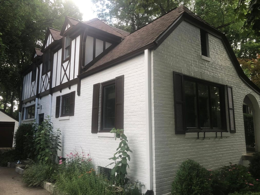 This is an image from Honeybear Painting of Mundelein, Inc. company, located in Mundelein, IL The image is a freshly painted white brick Tudor-style home with dark brown trim and shutters by Honeybear Painting of Mundelein