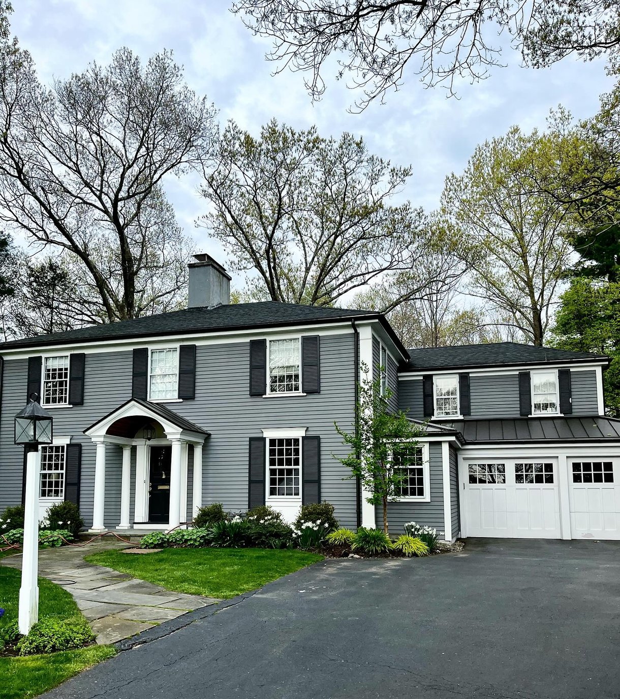 This is an image from TLC Painting Inc, located in Massachusetts. The exterior shows a classic two-story gray house with white trim and black shutters, freshly painted for a refined, modern look.