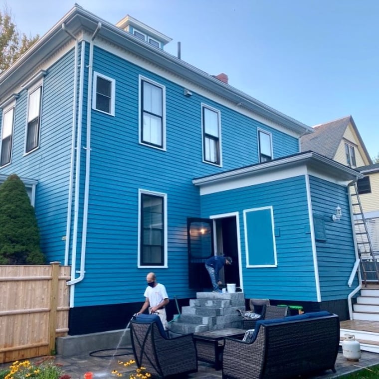 This is an image from Oliveira Painting Inc., located in Massachusetts. The photo shows a freshly painted two-story house with bright blue siding, white trim, and black accents, with workers completing the exterior project.
