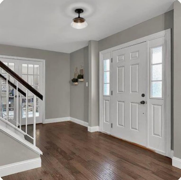 This is an image from Excel Painting company, located in Holly, MI The image is a freshly painted entryway with light gray walls, white trim, white front door with sidelights, hardwood floors, and staircase with white railing and dark wood handrail.