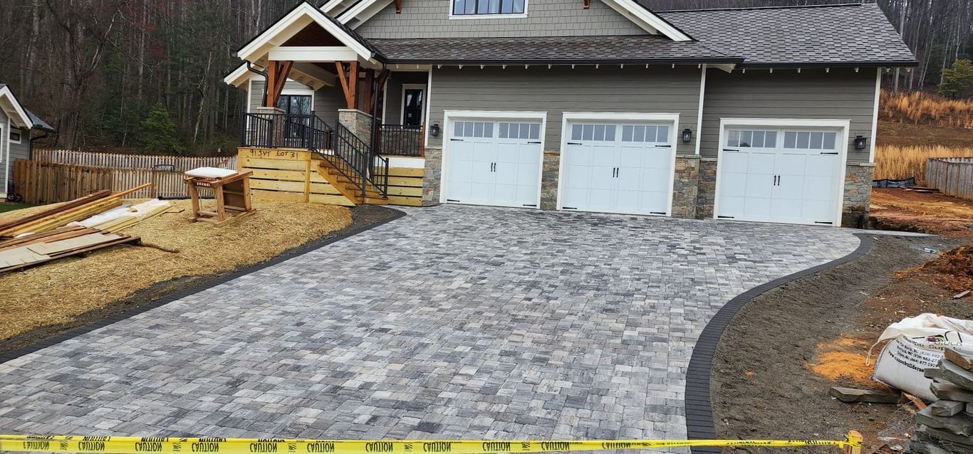 This is an image from JV Painting Services, located in North Carolina. This is a newly constructed driveway made of gray interlocking pavers leading to a three-car garage with white doors, in front of a modern suburban home.