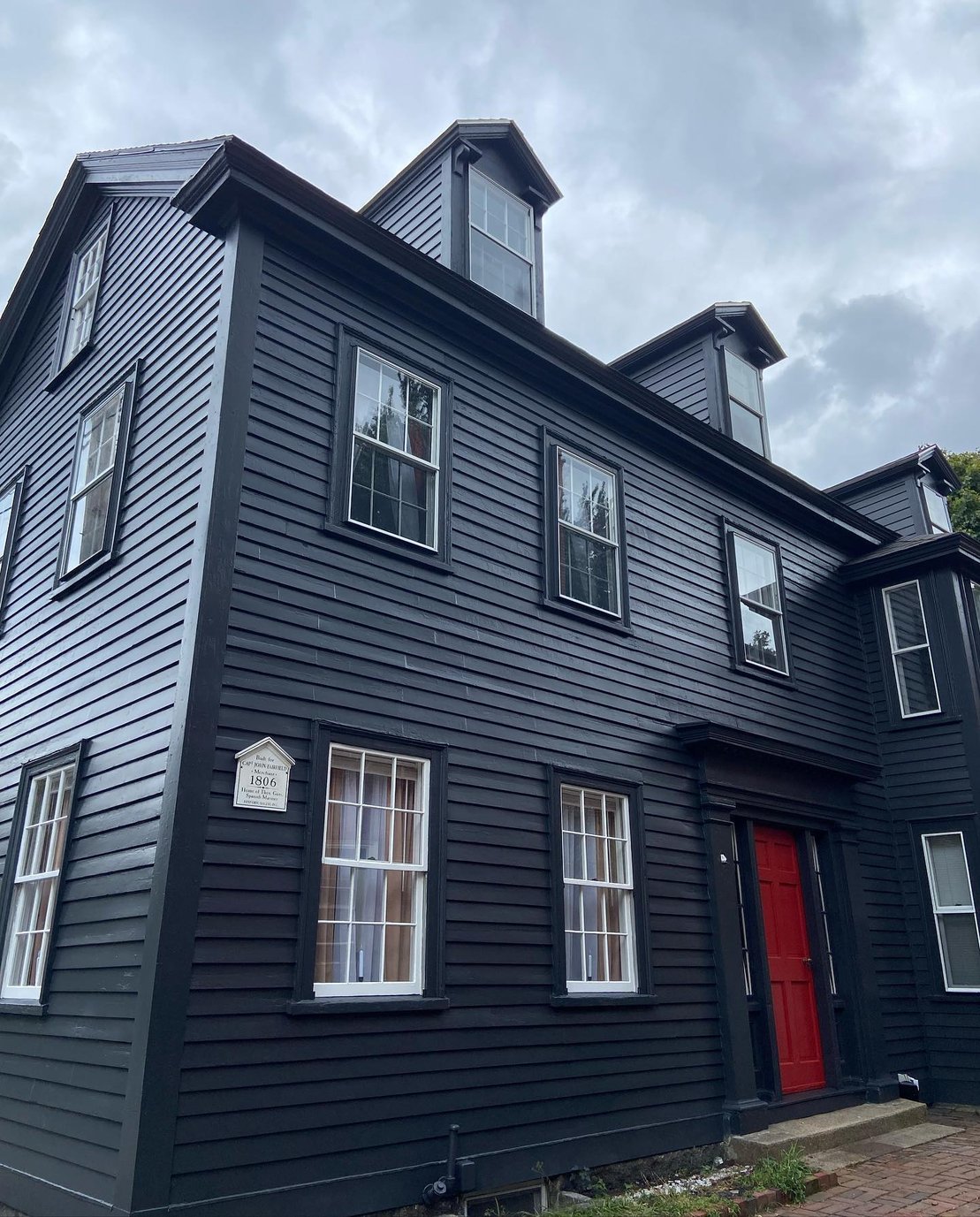 This is an image from BLC Painting, located in Massachusetts. The exterior of a historic dark navy home with a red front door and white-trimmed windows showcases a professional paint job under an overcast sky.