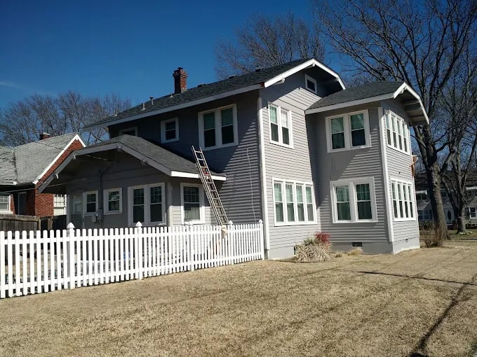 This is an image from John's Paint & Drywall, located in Oklahoma. The photo shows a freshly painted two-story home with gray siding, white trim, and a white picket fence under a clear blue sky.
