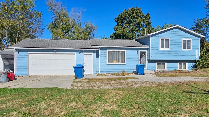 This is an image from Professional Painters Of Michigan company, located in Fowlerville, MI The image is a freshly painted light blue split-level house with white trim, attached garage, and clear blue sky, completed by Professional Painters of Michigan.