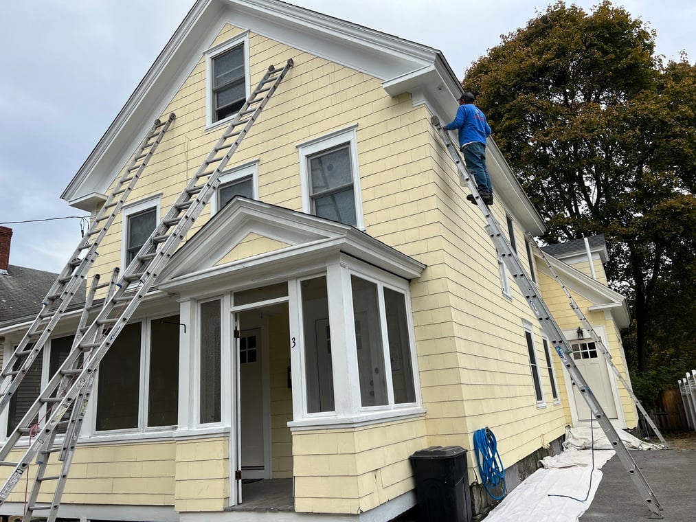 A painter works on the exterior of a light yellow house using ladders, preparing and painting the upper trim and siding.