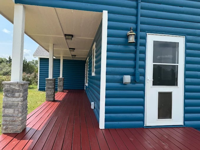 This is an image from CertaPro Painters of OKC, located in Oklahoma. The photo shows a freshly painted home exterior with bright blue log-style siding, white trim, and a red wooden porch floor with stone column bases.